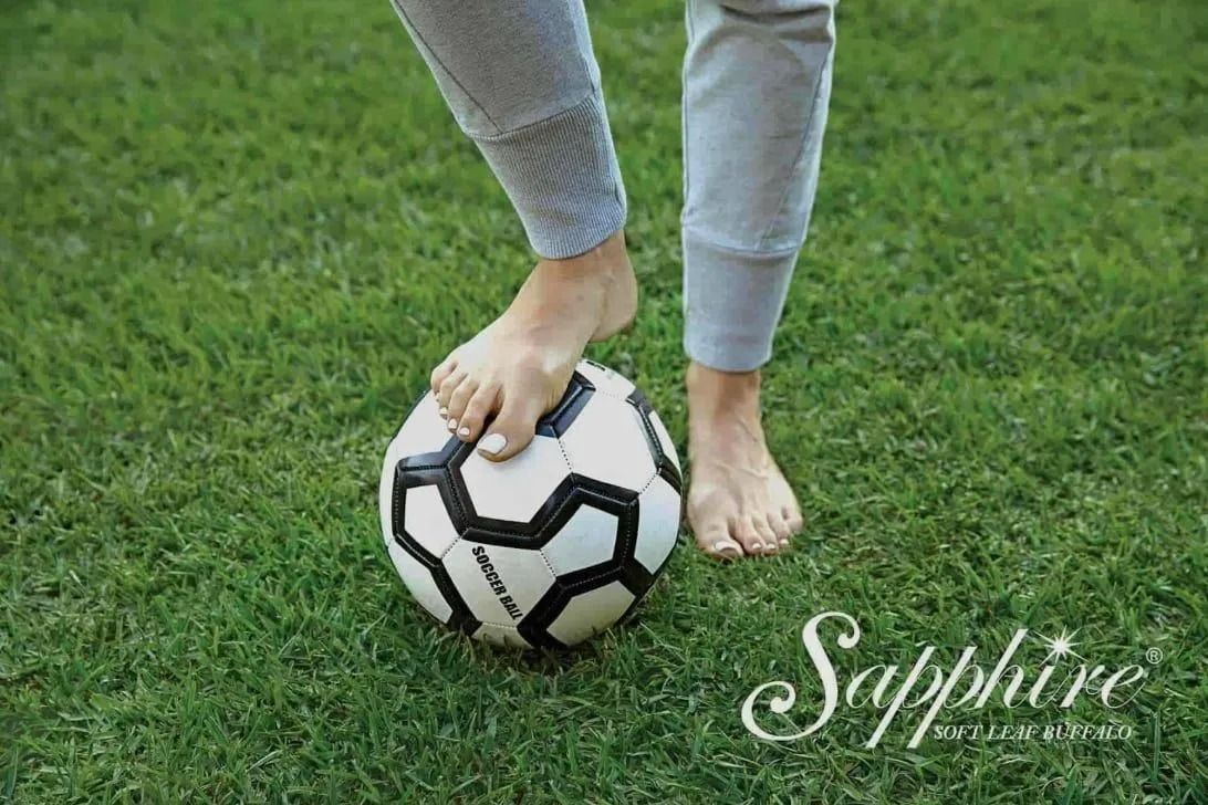 Bare feet on a soccer ball in grass. Person in gray sweatpants is standing on the ball, with a Sapphire logo in the corner.