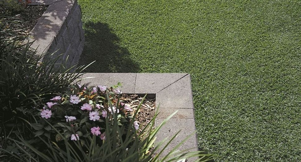 A stone planter box with pink flowers and green plants in front of a grassy lawn.