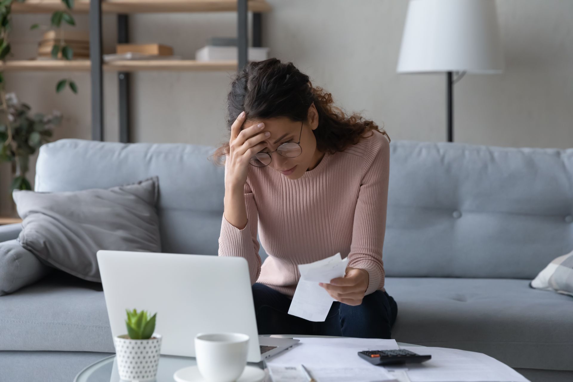 Distressed person at a laptop on a sofa, holding their head and reading a bill on the table.