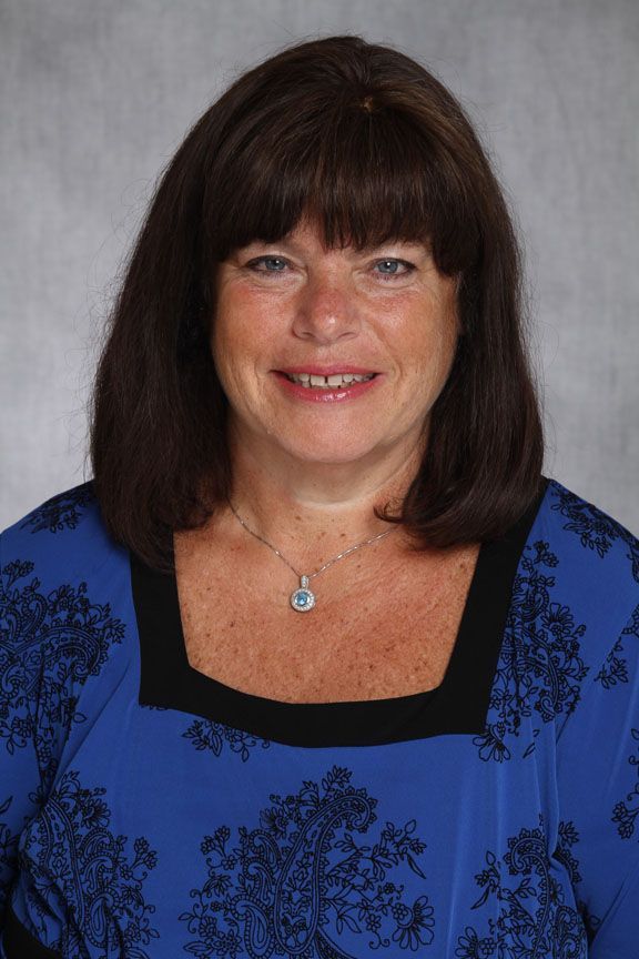 Woman with dark hair smiles, wearing a blue top with black trim, a necklace, against a grey background.