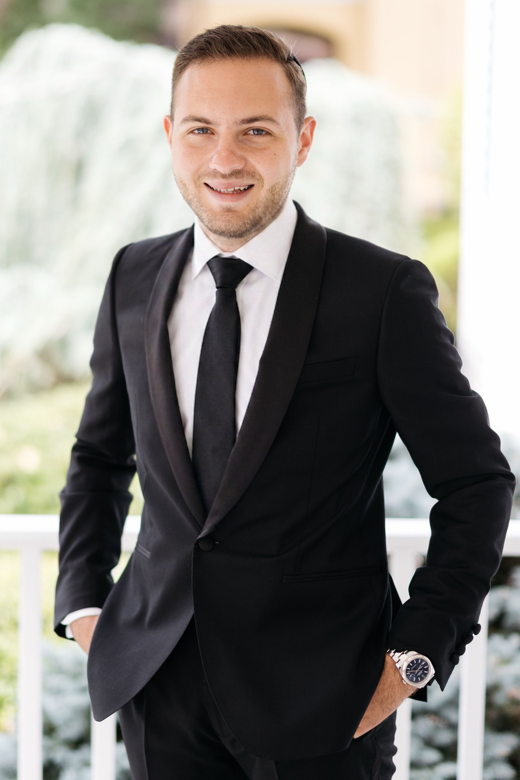 Man in a black suit and tie posing outdoors with a blurred bright background