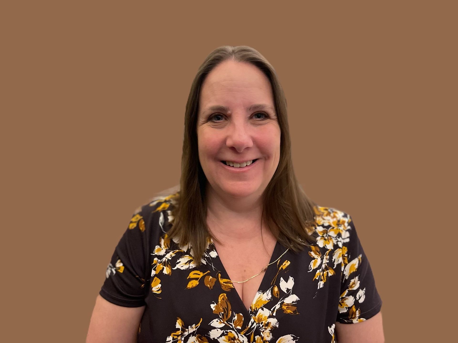 Woman smiling, wearing a floral print shirt, brown hair, against a brown backdrop.