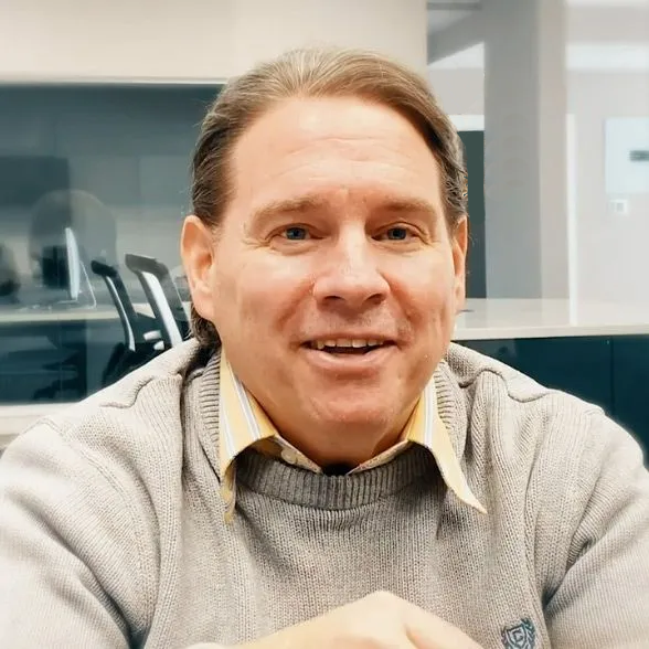 Man with light skin, smiling, wearing gray sweater over yellow collared shirt, in office setting.