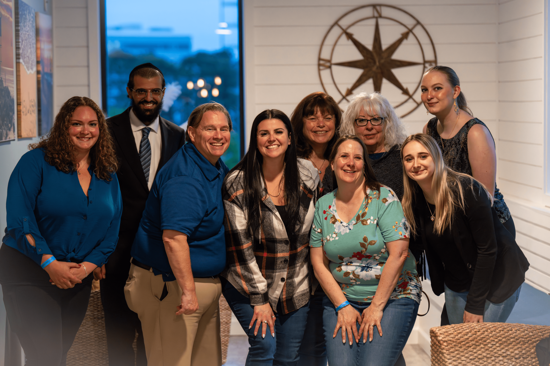 Group of nine people smiling indoors, with a nautical compass on the wall.