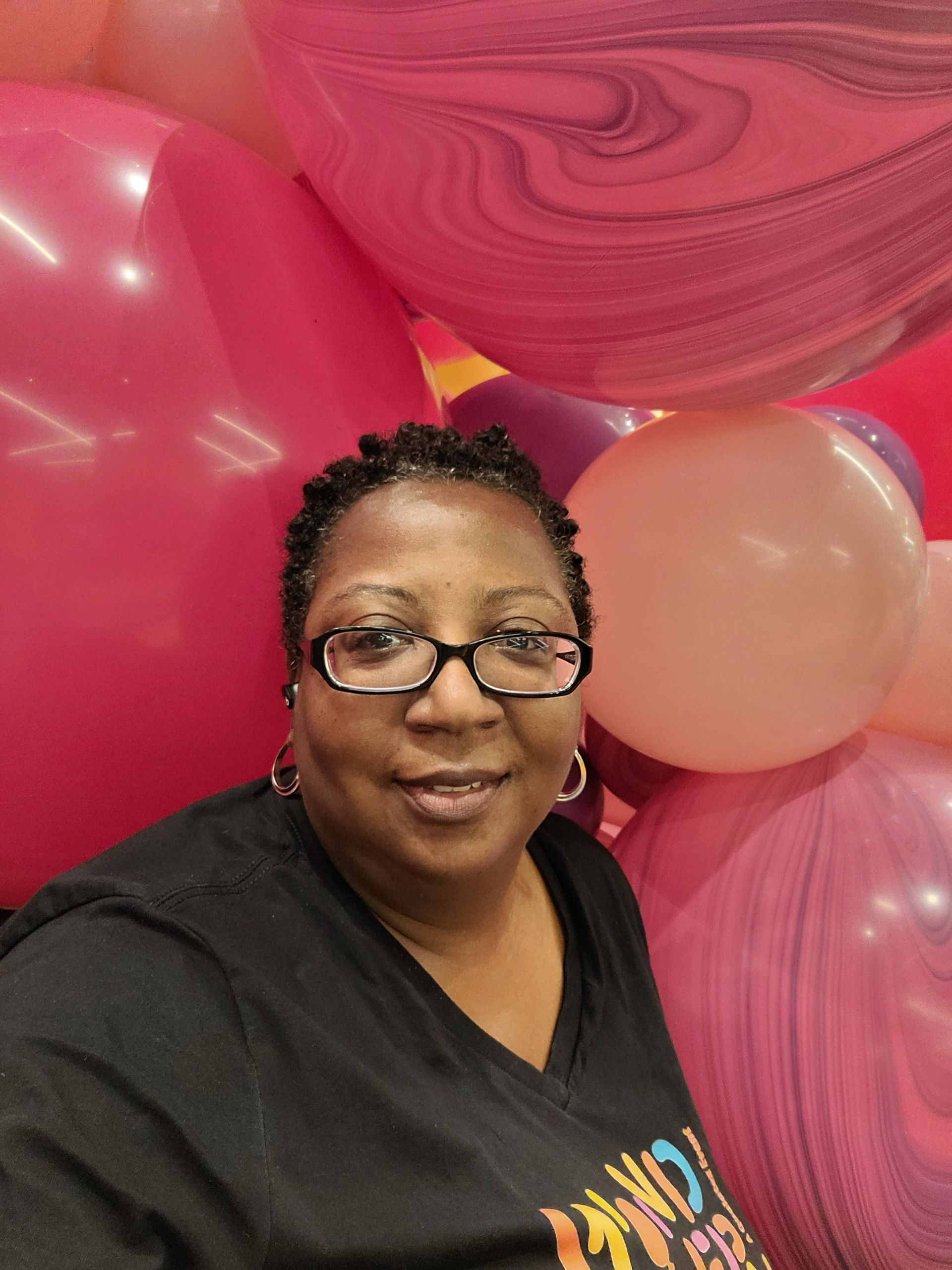 a woman wearing glasses is standing in front of a wall of pink balloons .