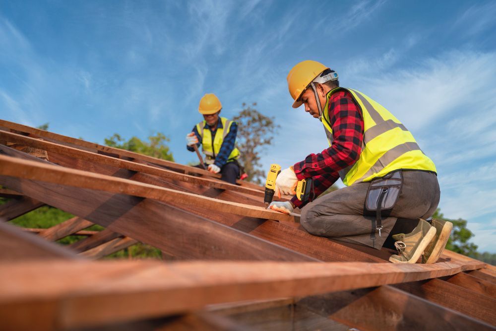 Two construction workers in hard hats and safety vests installing roof beams outdoors