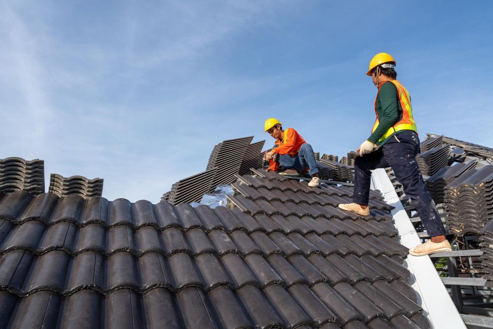 Roofers installing dark gray tiles on a sloped roof under a blue sky