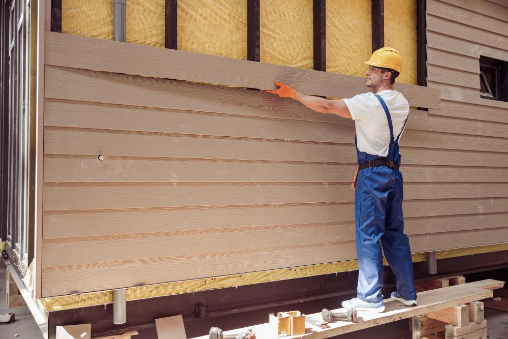 Worker in hard hat painting beige siding on a house exterior.
