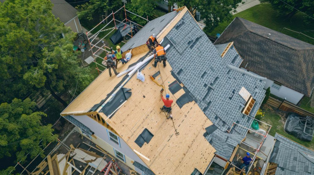 Workers repairing a roof with plywood, tarp, and shingles on a house surrounded by trees.