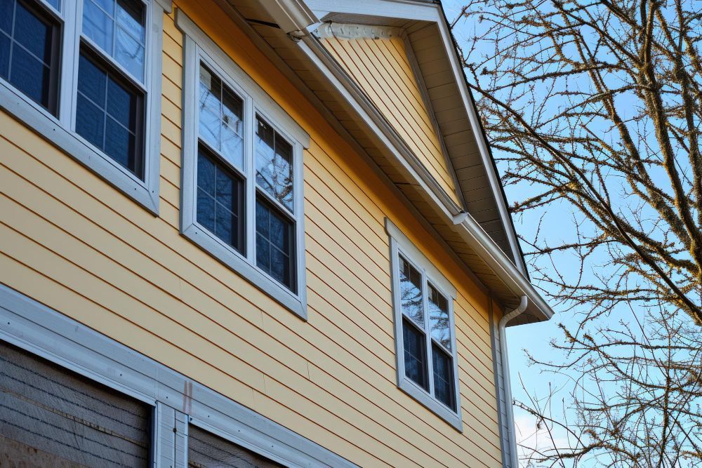 Yellow house siding with white-trimmed windows and bare tree branches against a blue sky