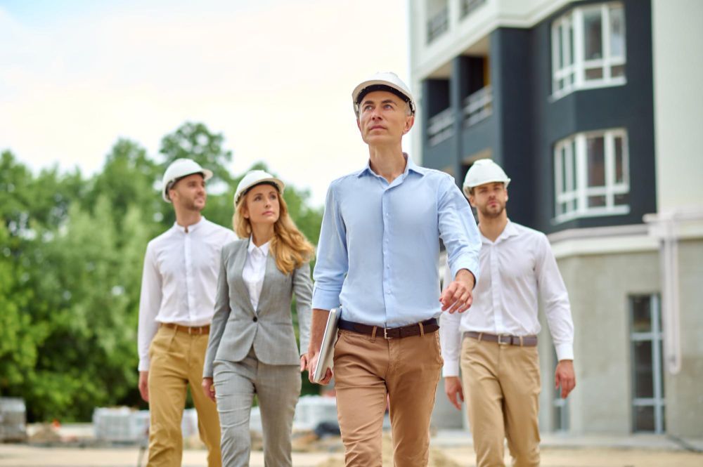 Four coworkers in hard hats walking outside a modern building, one in front leading.