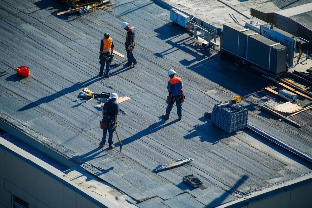Construction workers on a rooftop, wearing orange vests and hard hats, with materials and equipment nearby