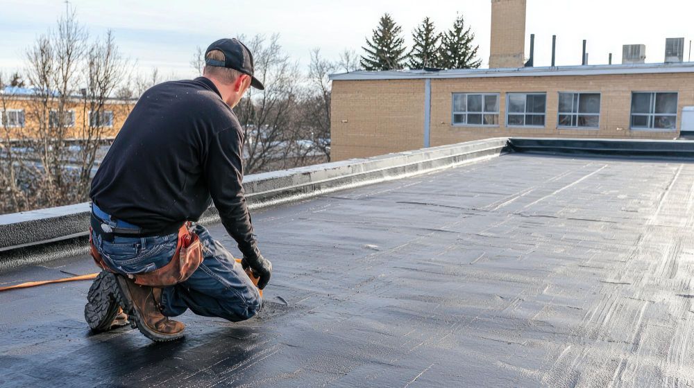 Worker kneeling on a flat rooftop, applying black roofing material near a building