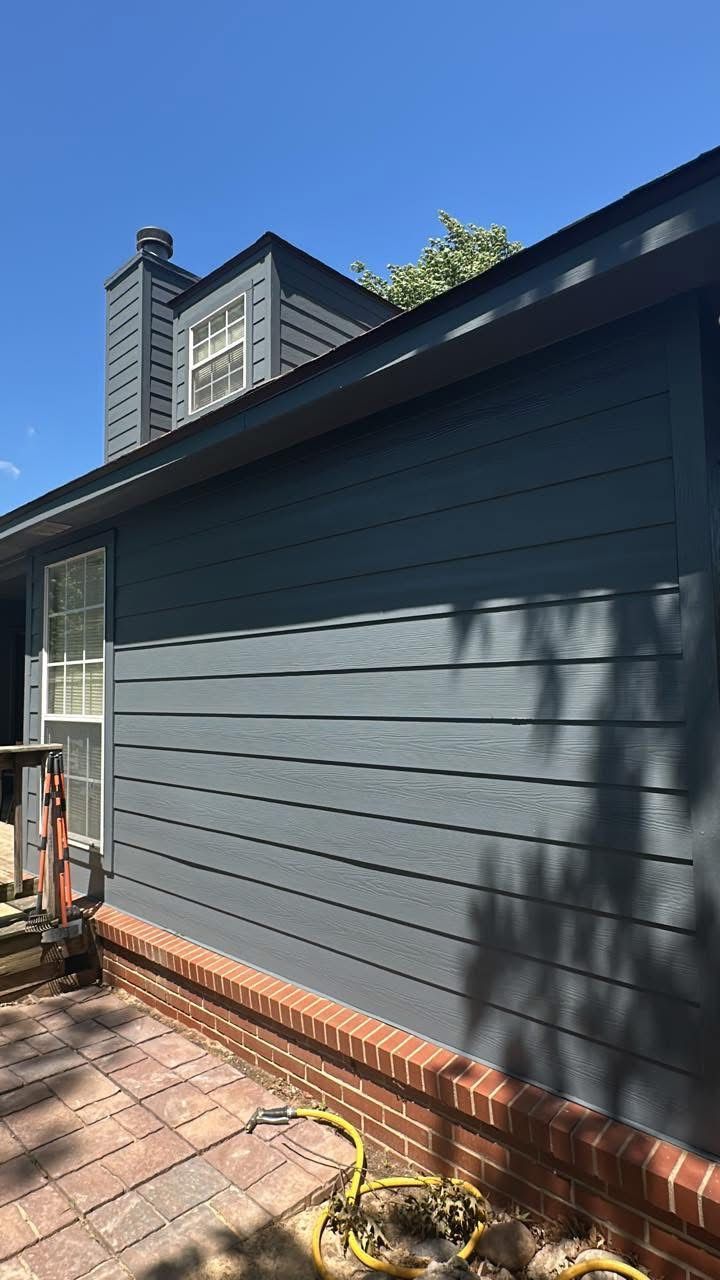 Dark gray house exterior with horizontal siding, brick foundation, and a tall window under a blue sky.