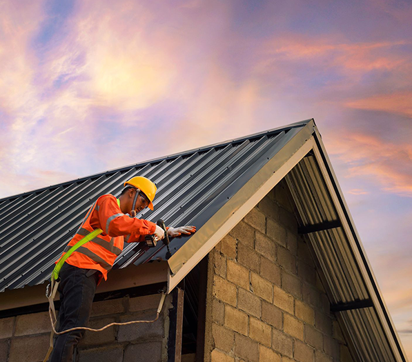 Worker in orange safety gear and yellow hard hat installing roofing on a house at sunset
