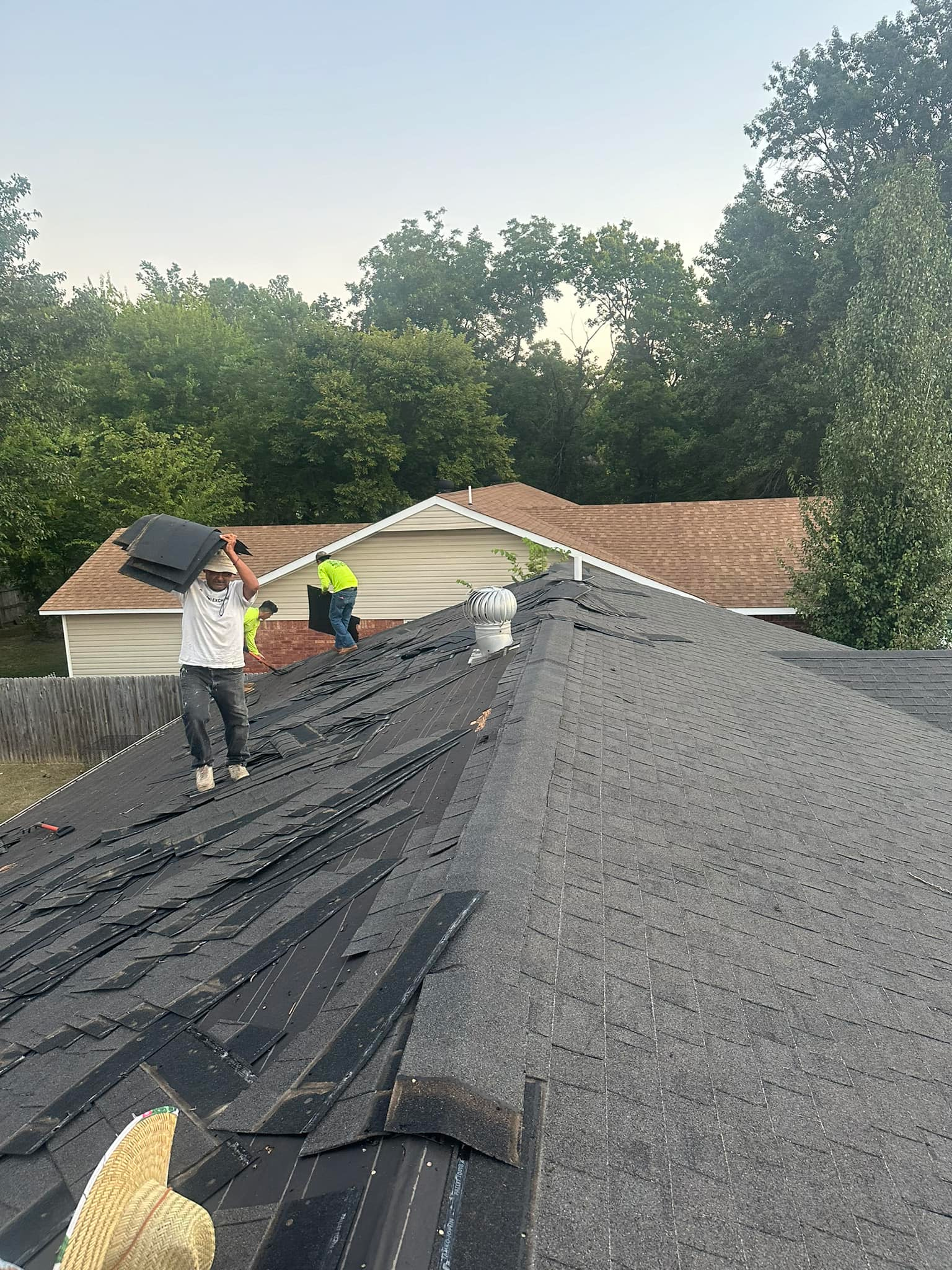 Roofers working on a shingled roof under a clear sky, with one worker on the slope and another near the ridge.