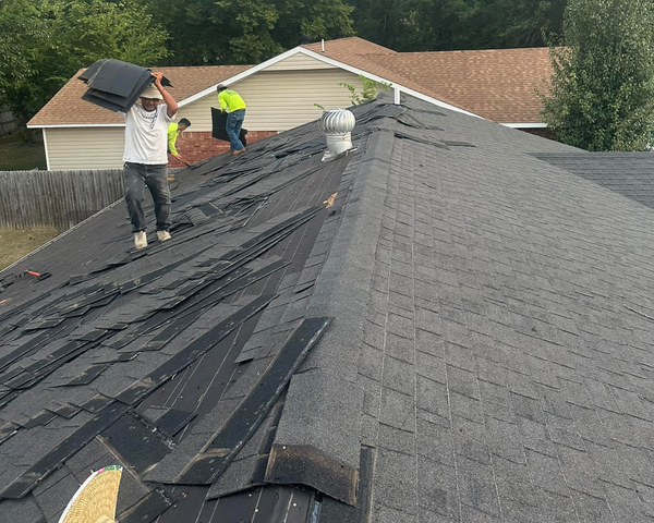 Workers repairing a dark shingle roof on a house, with one lifting underlayment near the ridge.
