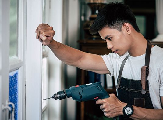 Person using a power drill to install a white window frame indoors