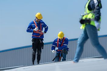 Workers in hard hats and safety harnesses on a rooftop, one carrying equipment and one kneeling.