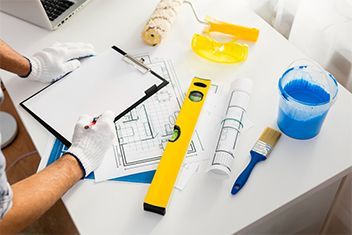 Person drawing plans at a desk with a yellow level, blue tape, and paint bucket