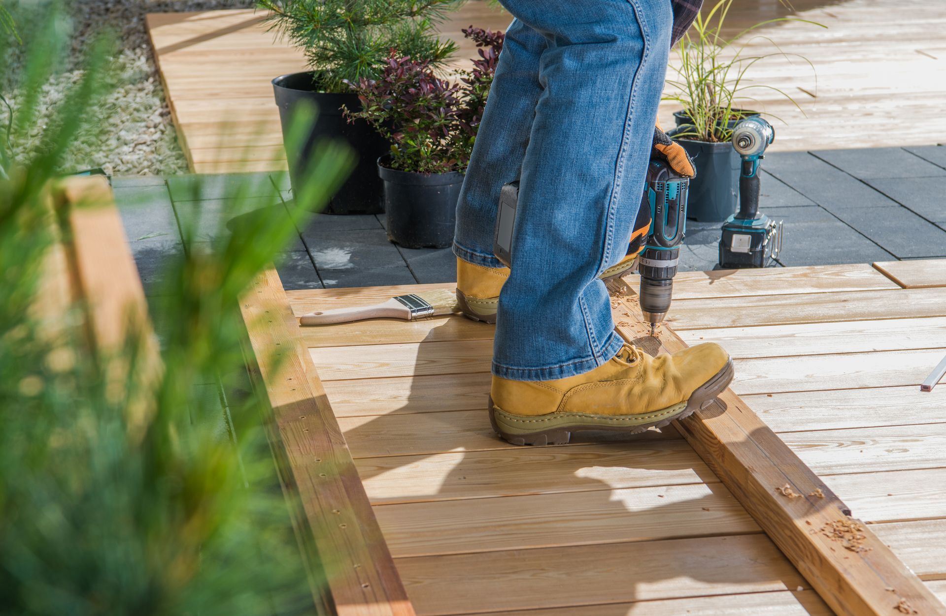 Person in jeans and tan boots standing on a wooden deck beside plants and tools.