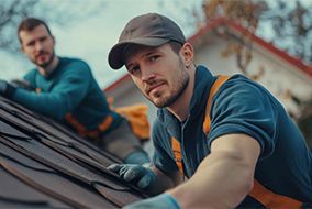Two roofers in work clothes repairing a roof, one in focus in the foreground.