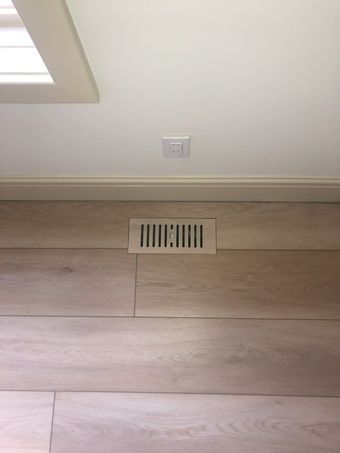 Black floor vent on light wood flooring beside a white baseboard.