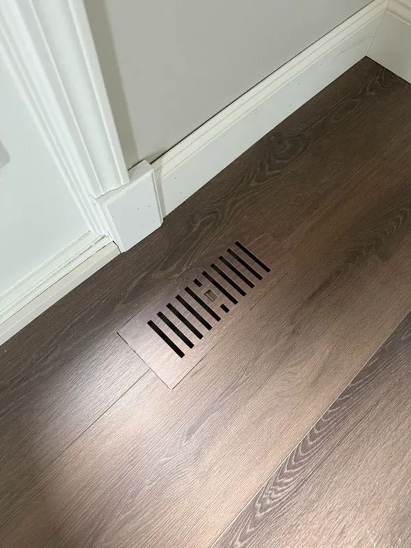 Floor vent in a dark wood floor near a white wall and baseboard.