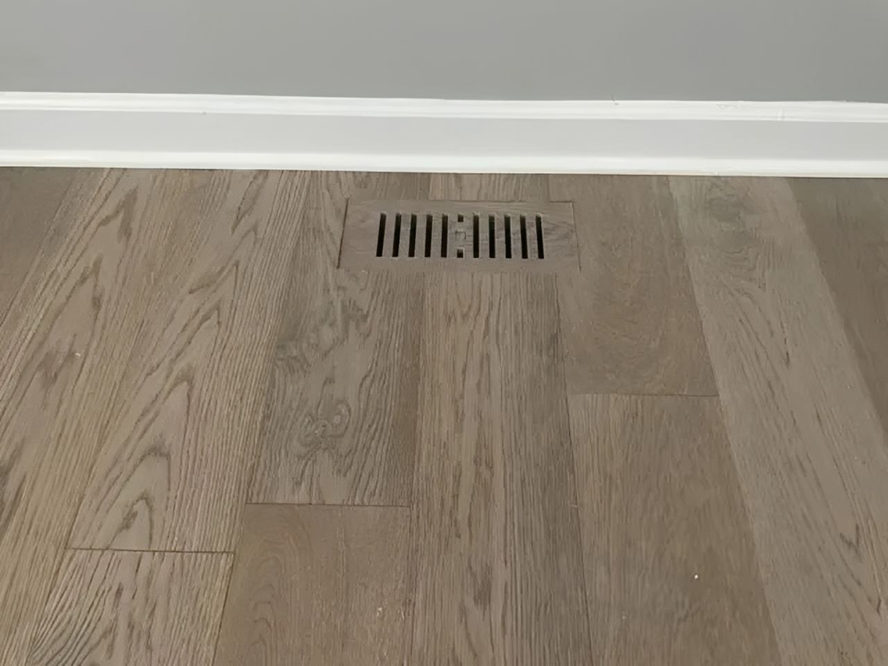 Wood floor with a rectangular vent beneath a white baseboard
