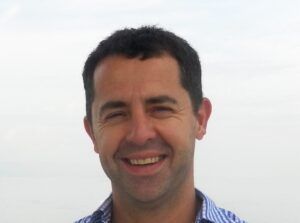 Smiling man outdoors against a bright sky, wearing a blue checkered shirt