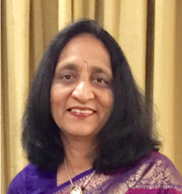 Portrait of a smiling woman in a purple sari, standing before beige curtains.