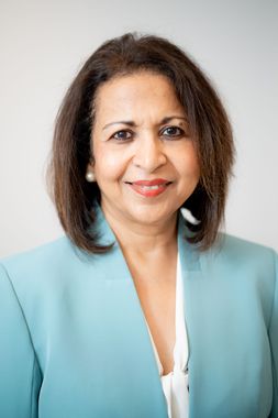 Headshot of a smiling person in a navy blazer and necklace against a light gray background