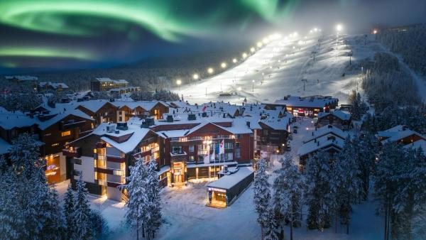 Snowy mountain village at night with glowing cabins and ski slope under green aurora lights