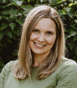 Smiling blonde woman in a green shirt outdoors with leafy greenery behind her