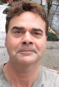 Close-up selfie of a man outdoors on a sidewalk, wearing a light gray shirt with a neutral expression.