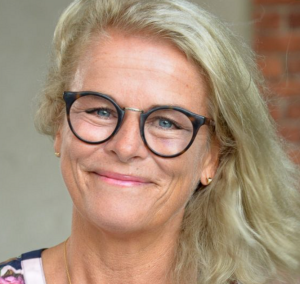 Smiling person with blonde hair and black glasses against a plain background
