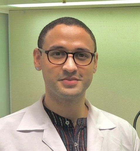 Person in glasses and a white lab coat standing against a green background.