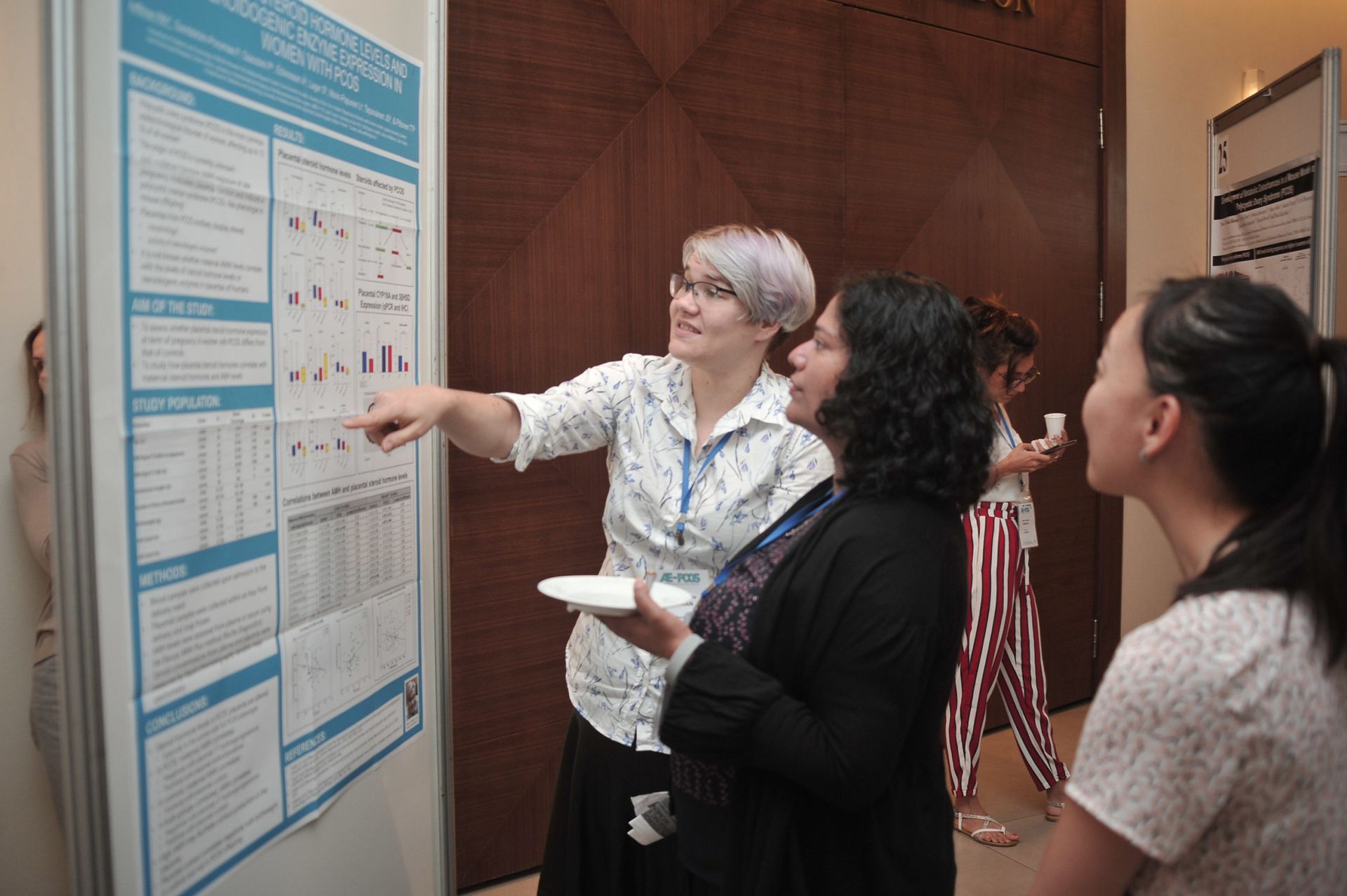Three people examine a large blue poster board indoors, one pointing at charts and notes.