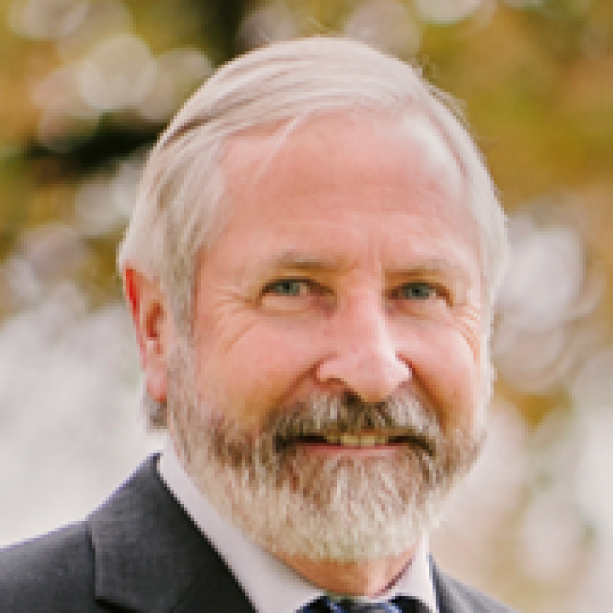 Portrait of a smiling man in a dark suit with gray hair and beard against a blurred outdoor background