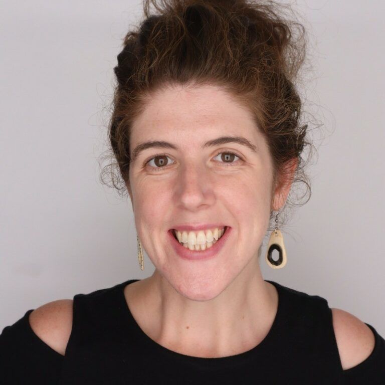 Smiling person in a black top with cutout shoulders, wearing dangling earrings against a plain background
