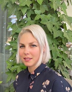 Woman with short blonde hair in a dark floral blouse, standing beside leafy green vines outdoors
