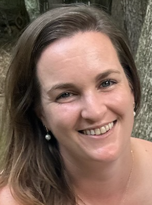 Smiling woman with brown hair in a dark top, standing outdoors by a tree.