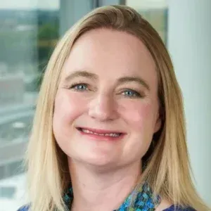 Smiling woman with blonde hair in a blue patterned blouse, in a bright office setting