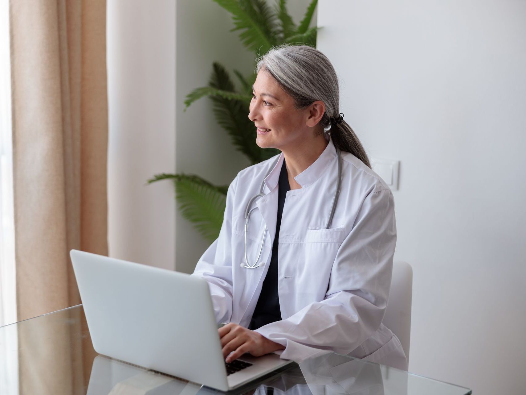 A female doctor is sitting at a desk using a laptop computer.