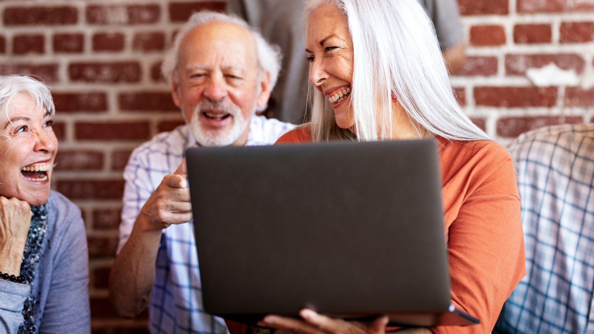 Three seniors smiling and chatting around a laptop in a brick-walled room
