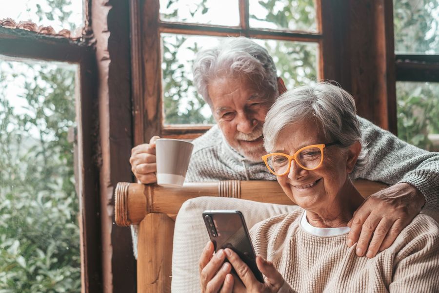 Older couple smiling while looking at a phone by a window, one holding a mug