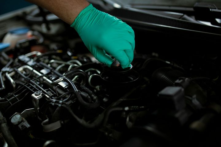 A hand in a green glove reaches into a dark car engine bay to perform maintenance.