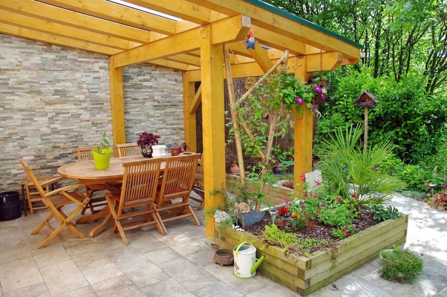 Wooden patio dining area beside a stone wall, with potted flowers and a garden pergola.