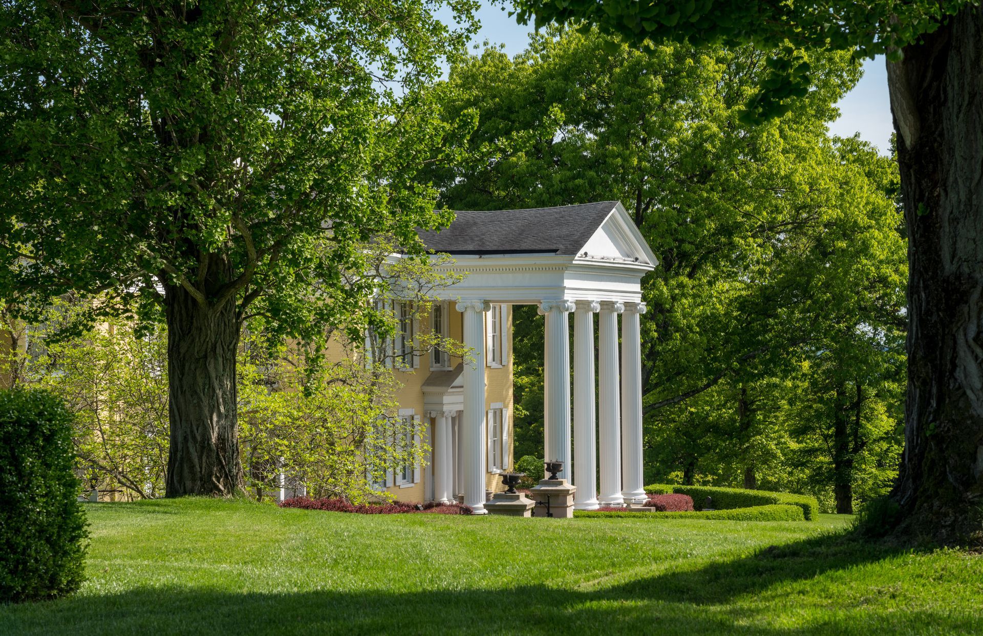 Small neoclassical pavilion with white columns on a grassy lawn, surrounded by trees.