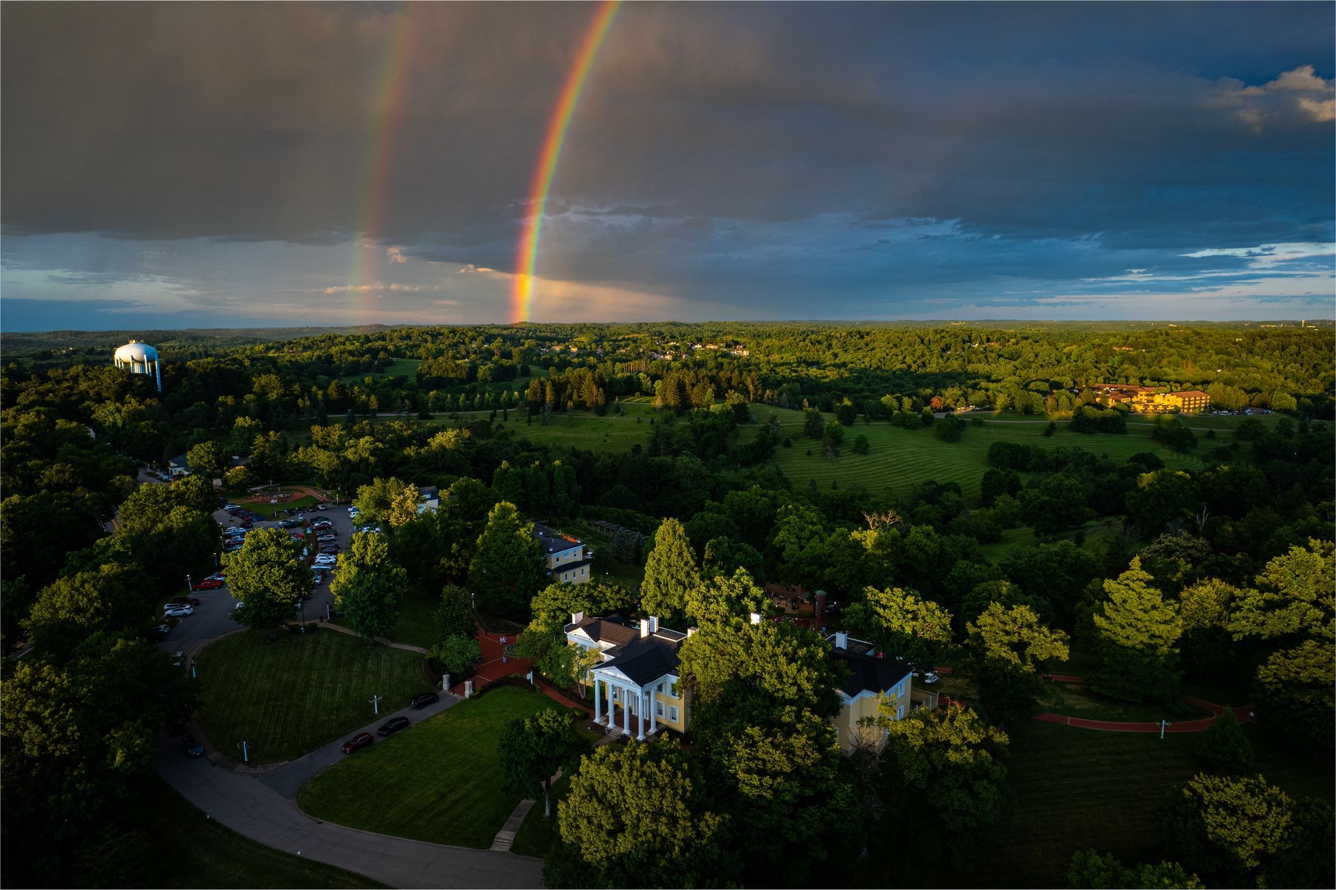 Double rainbow arches over a lush green landscape, with a white-columned house in the foreground.